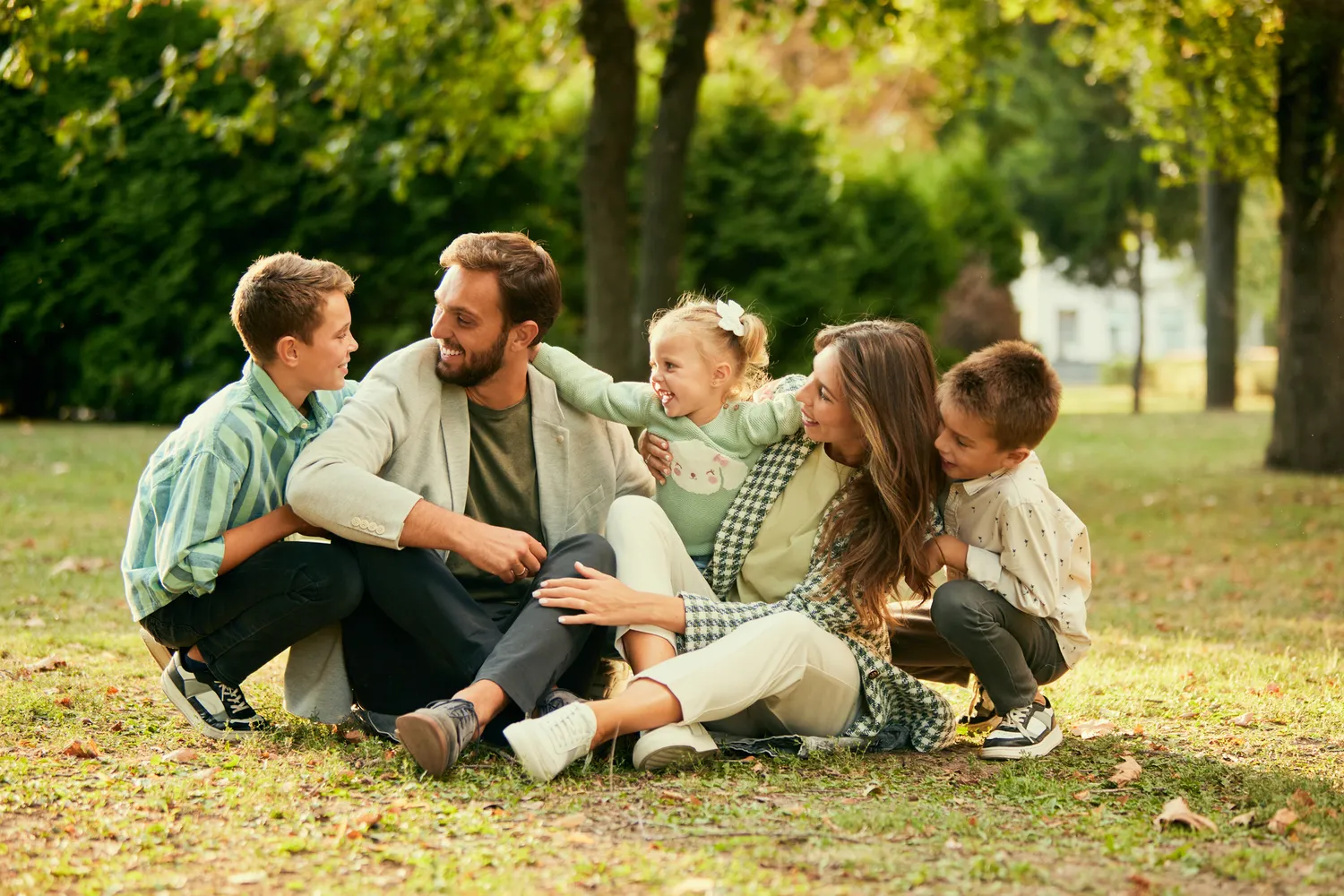 Herzensnah by Tanita Englisch - Familiäre Unterstützung - Eine große Familie, bestehend aus zwei Erwachsenen und vier Kindern, sitzt zusammen auf der Wiese in einem Park, lächelt und unterhält sich miteinander.