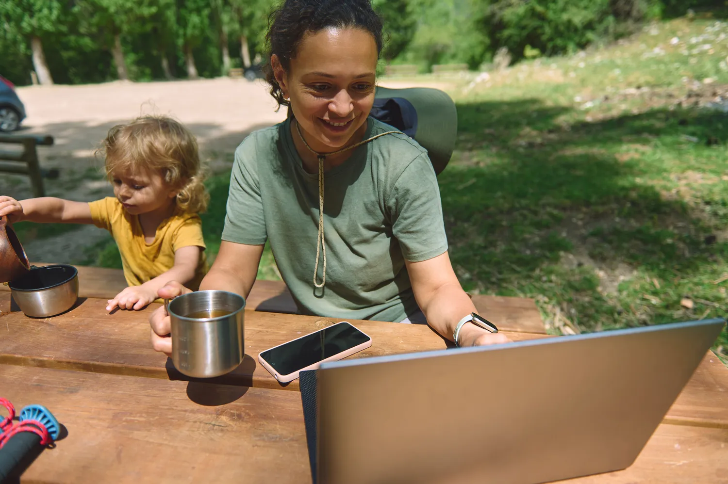 Herzensnah by Tanita Englisch - Familiäre Unterstützung - Eine Frau und ein kleines Kind sitzen im Freien an einem Holztisch und schauen beide auf einen Laptop-Bildschirm. Ein Smartphone und ein Metallbecher stehen ebenfalls auf dem Tisch.